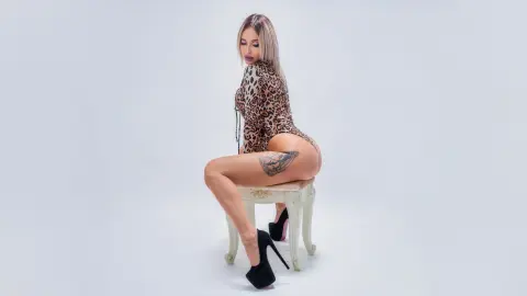 Blonde woman in a leopard print bodysuit and black high heels sitting on a white stool against a plain background.