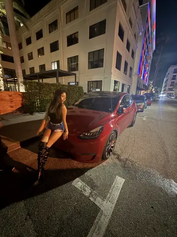 Stylish woman in denim shorts and tall black boots posing by a red Tesla on a city street at night.