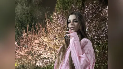 Young woman in a pink long-sleeve shirt posing in front of blooming pink flowers at night.
