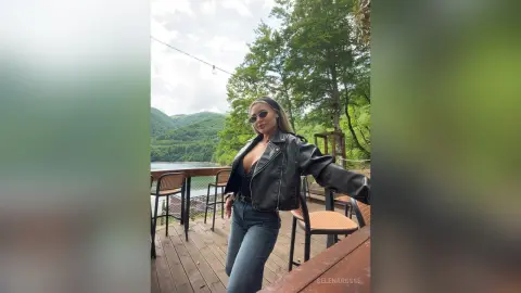 SelenaRusse poses on an outdoor deck wearing a black leather jacket, a low-cut black top, and blue jeans.