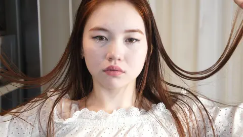 Young woman with long brown hair wearing a white eyelet blouse in a bright, minimalist indoor setting.
