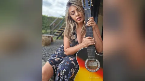 CrisstyBaez — CrisstyBaez sits outdoors in a floral print dress while holding an acoustic guitar.