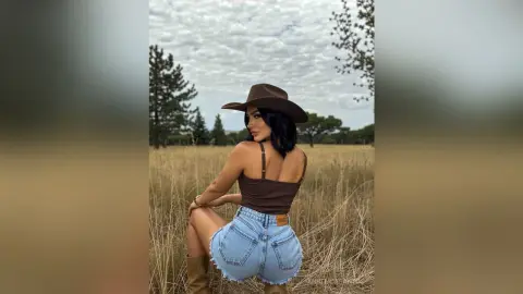 Stylish woman in a brown tank top and denim shorts wearing a cowboy hat in a golden field under a cloudy sky.