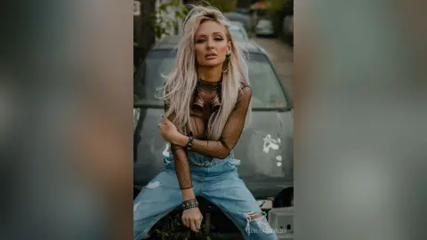 LeslyAnderson poses sitting on a dark car wearing a black mesh long-sleeve top and light blue denim jeans.