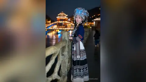 Young woman in traditional ethnic costume and silver headpiece standing by a lit bridge at night.