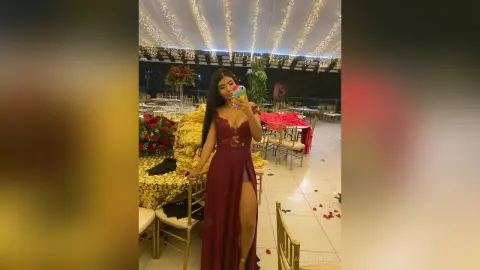 Woman in elegant burgundy evening gown posing in a decorated banquet hall with fairy lights and floral decor.