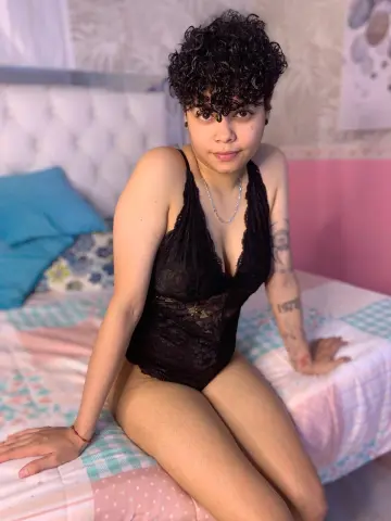 Young woman in black lace bodysuit sitting on a bed in a bright, cozy bedroom setting.