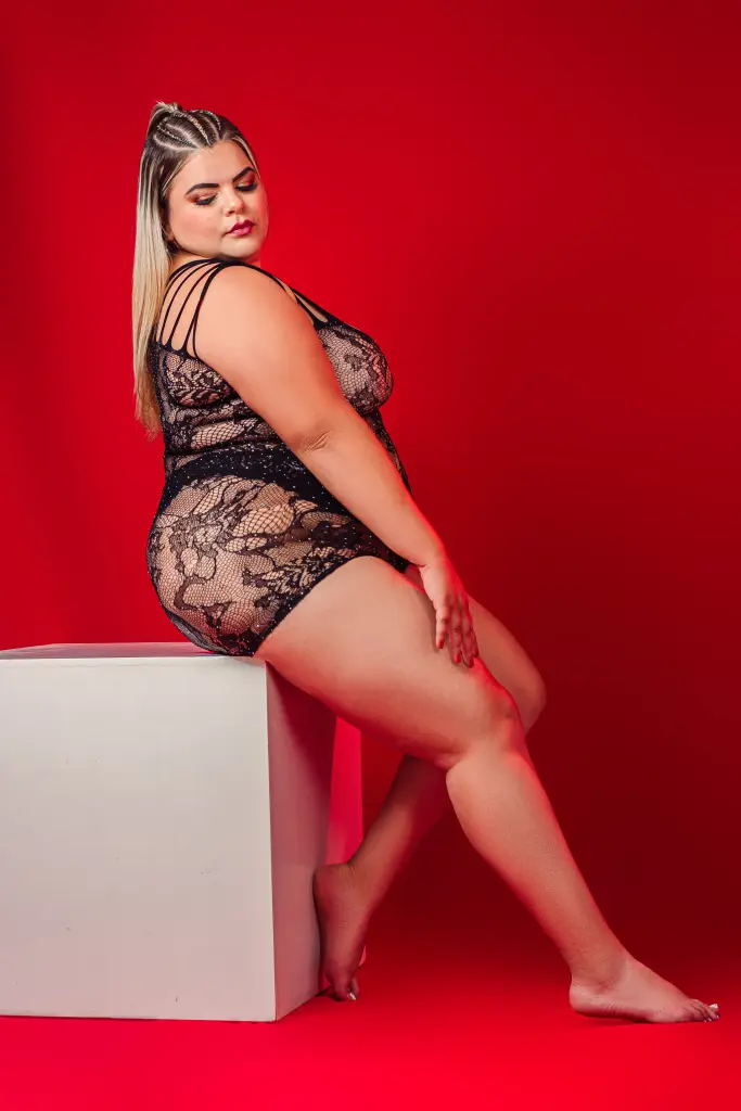 Smiling blonde woman in a black bodysuit relaxing on a white background for a confident lifestyle portrait.