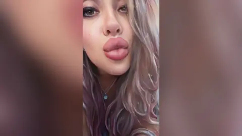 Close-up portrait of a young woman with long wavy hair and pink lipstick posing with a playful pouty expression.
