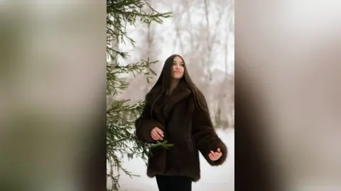 Young woman in a cozy brown faux fur coat standing in a snowy winter forest setting.
