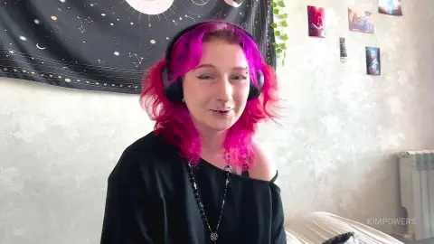 Smiling woman with bright pink hair and black outfit wearing headphones in a cozy bedroom with celestial decor.