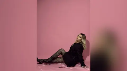 Elegant woman in a black dress and patterned tights posing against a soft pink background.