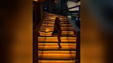 TefyRusso sits on a lit staircase wearing a black bra and black panties, posing with her hands near her face.