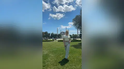 Blonde woman in a cream crop top and white trousers standing on a sunny green lawn near a mosque.