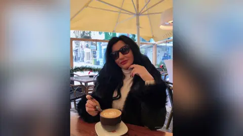 MissyJolie sits at an outdoor cafe table wearing sunglasses and a black furry coat, holding a spoon over a cup of latte foam.