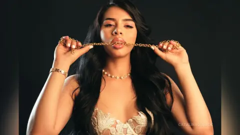 CatalinaCusack bites a gold chain held between her teeth while wearing a white lace bra and a pearl necklace.