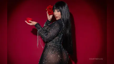 Glamorous woman in a sheer black sequined dress holding red roses against a moody red background.