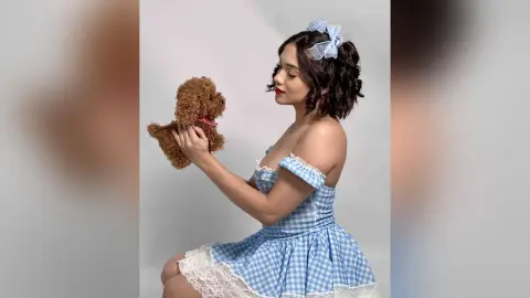 Young woman in a blue gingham lace dress holding a brown teddy bear against a white background.