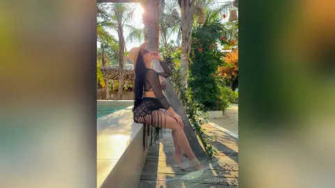 NicoleBella poses outdoors by a pool wearing a black lace bra and matching fringe skirt with a tan sun hat.