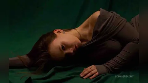 SophiePeachy lies on a green sheet wearing a brown long-sleeve top, looking toward the camera with her hair spread out.