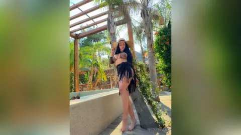 NicoleBella stands outdoors in a black fringe bikini and a wide-brimmed straw hat, posing next to a stone wall and palm trees.