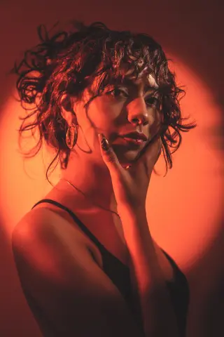 Moody portrait of a woman with curly hair in a black camisole under warm red lighting.