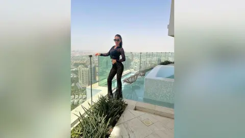 ArianaKroft stands on a high-rise balcony wearing a black long-sleeved crop top and black lace leggings, overlooking a city skyline.