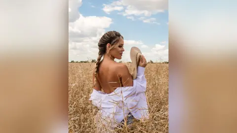 Blonde woman in a white linen shirt and sun hat standing in a golden wheat field under a blue sky.