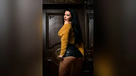 EllaHadid poses in a yellow fuzzy sweater and black leather mini skirt, looking back over her shoulder with long dark hair.