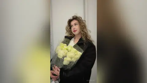 Blonde woman in a black coat holding a bouquet of yellow flowers in a bright indoor setting.