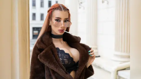 DeviNessa wears a brown fur coat and black lace bra while holding a cigarette in her mouth against a white pillared background.