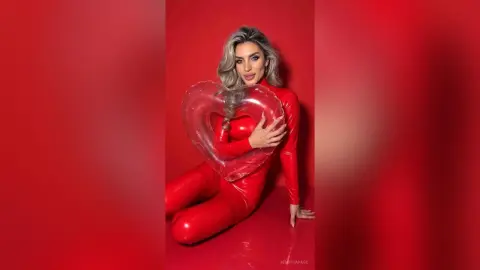 JessycaPage poses in a tight red bodysuit while holding a large red heart balloon against a red background.
