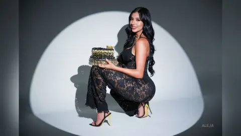 Aleja poses in a black lace jumpsuit and high heels, holding a decorative black and gold tray against a white circular background.