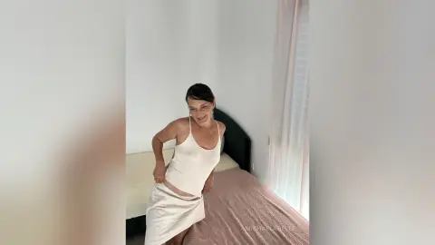 MichaelaBelle smiles while wearing a white ribbed tank top and matching skirt, standing next to a bed.