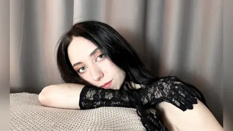 Young woman with long dark hair wearing a black lace top leaning on a textured beige pillow in a soft setting.
