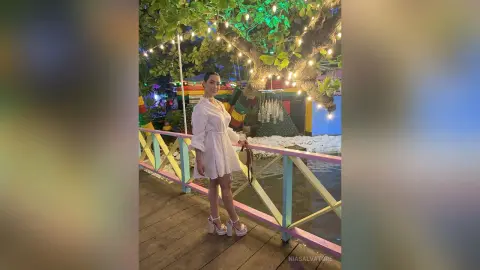 Elegant woman in a white dress posing on a wooden pier under festive string lights at a tropical night garden.