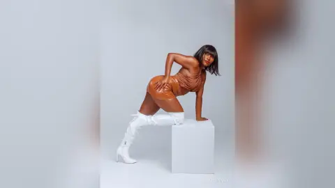 Confident woman in a brown leather mini dress and white tall boots posing against a white studio background.