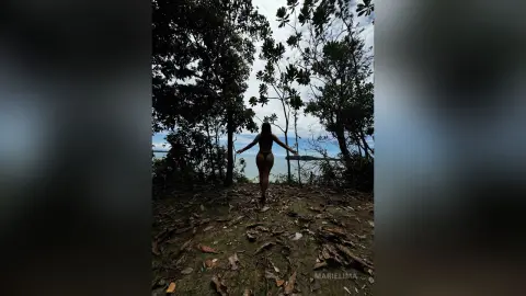 Back view of a woman in a bikini standing in a lush tropical forest overlooking the ocean at dusk.