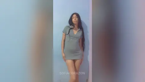 SofiaCorazon poses against a light blue wall wearing a tight, short grey dress that shows off her long dark hair and legs.