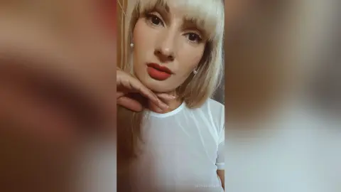 Blonde woman in a white mesh top with red lipstick posing with a serene expression indoors.
