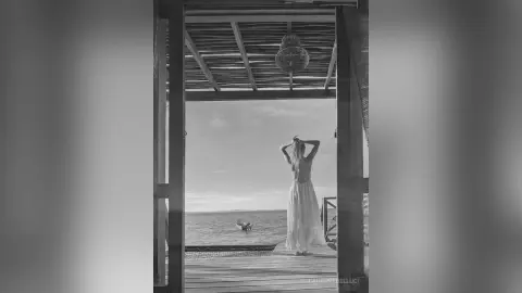 LaurentBelluci stands facing the ocean, wearing a long white dress and holding her hands behind her head.