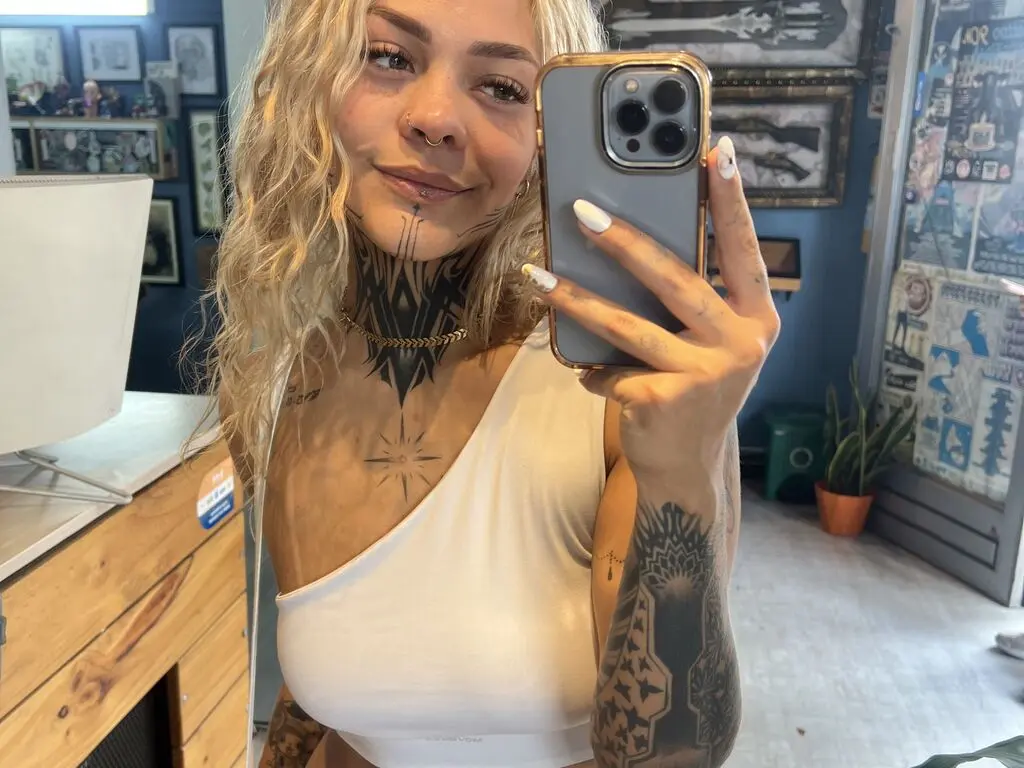 Tattooed woman in a white tank top taking a selfie in a room filled with framed art and decor.