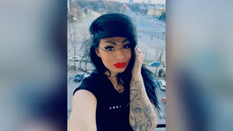 DeeaAnderson poses wearing a black t-shirt and red lipstick with her long dark hair styled over one eye against a city street background.