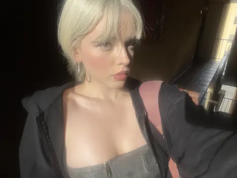 Blonde woman in a black jacket and grey corset posing in warm, cinematic sunlight.