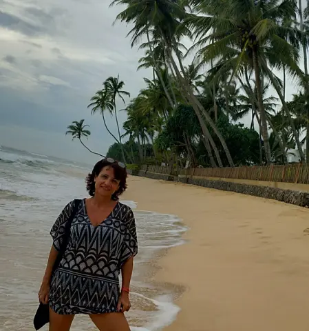 Smiling woman in a patterned black and white beach cover-up standing on a tropical sandy beach by palm trees.