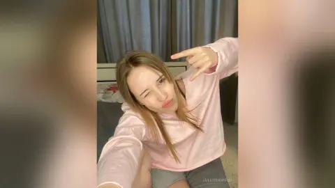 Young woman in a pink sweatshirt posing playfully in a cozy bedroom setting.