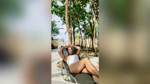 Young woman in a white bikini relaxing on a lounge chair in a sunny tropical garden setting.