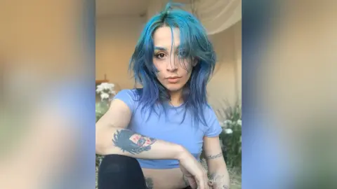 Woman with blue hair in a light blue crop top posing in a bright indoor setting with a relaxed mood.