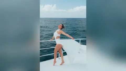 Elegant woman in a flowing white summer dress posing on a yacht deck against a blue ocean background.