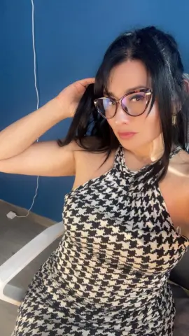Woman in a black and white houndstooth halter dress wearing glasses against a blue wall.